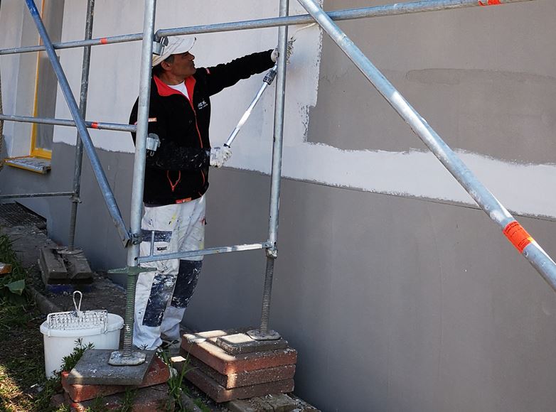 Ein Bauarbeiter trägt Putz auf eine Außenwand auf, während er auf einem Gerüst steht, ausgerüstet mit Eimer und Kelle. Der Arbeiter trägt einen weißen Overall und eine schwarze, mit nachhaltiger Isolierung gefütterte Jacke.
