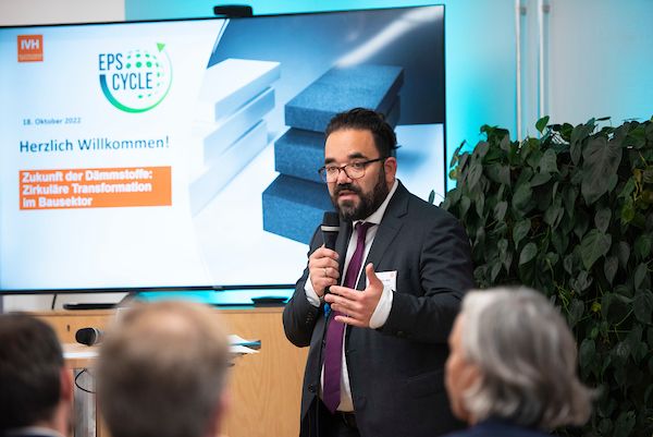 Ein Mann mit Bart, Brille und Anzug spricht an einem Podium mit Mikrofon vor einem Publikum. Auf einem Präsentationsbildschirm hinter ihm werden Texte und Logos zu einer Konferenz über Nach