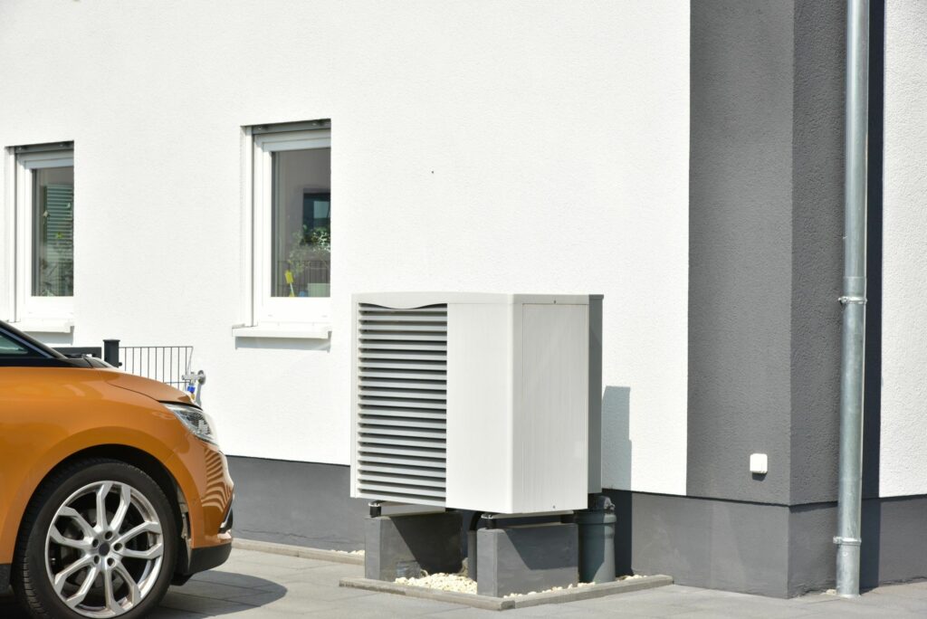 Eine Außenwärmepumpe, die neben einem Gebäude mit weißen und grauen Wänden unter klarem Himmel installiert ist. Im Vordergrund ist die vordere linke Seite eines orangefarbenen Autos zu sehen, das neben einer mit nachgedämmten Wand geparkt ist.