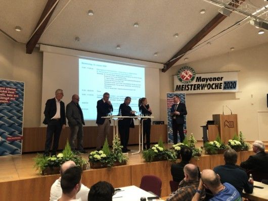 Eine Gruppe von Rednern steht während der „Mayener Meisterwoche 2020“ an einem Podium in einem Konferenzraum, die Teilnehmer sitzen ihnen gegenüber. Auf einer Projektionsleinwand wird ein Text über „n