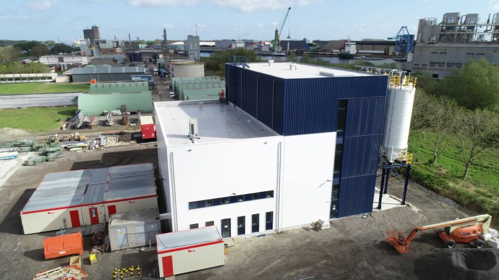 Luftaufnahme einer modernen Industrieanlage mit mehreren Gebäuden, darunter eine große weiß-blaue Struktur aus Styropor, umgeben von Baumaschinen und grünen Feldern.
