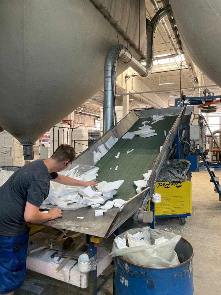 Ein Arbeiter sortiert Stücke aus weißem EPS-Hartschaum auf einem Förderband in einer großen Industriehalle. Darüber sind große Lagertanks zu sehen.