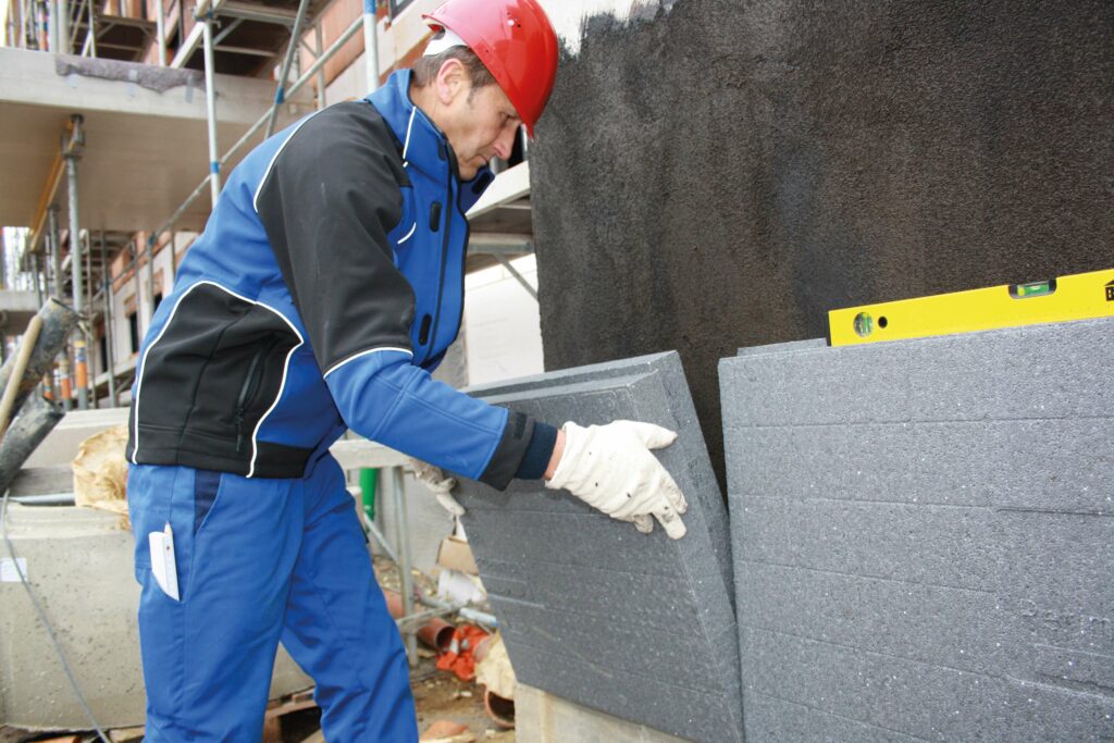 Ein Bauarbeiter in blauer Uniform und rotem Helm trägt auf einer Baustelle sorgfältig Mörtel zwischen großen grauen Styroporblöcken auf und verwendet dabei eine Wasserwaage, um die Ausrichtung sicherzustellen.