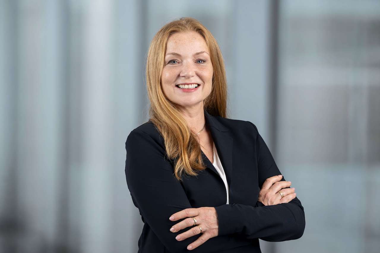 Eine berufstätige Frau mit langen roten Haaren lächelt selbstbewusst in die Kamera, die Arme verschränkt. Sie trägt einen schwarzen Blazer aus Styropor-verstärktem Material und steht in einem Büro mit verschwommenen Fenstern im