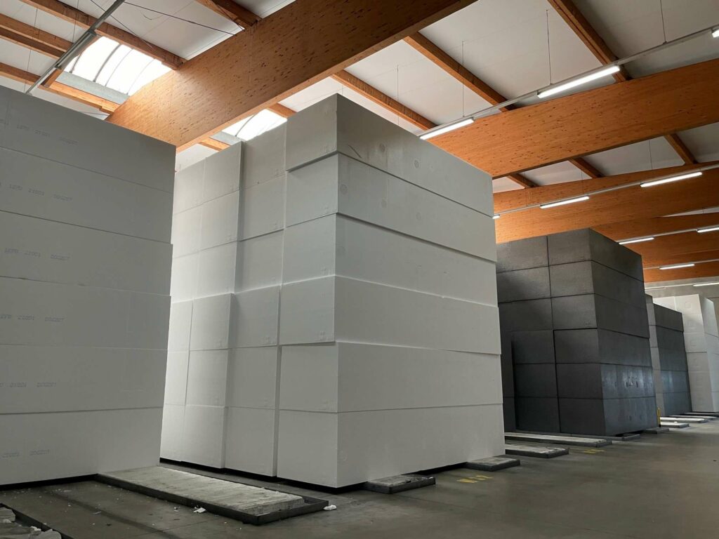 Eine große Lagerhalle mit freiliegenden Holzbalkendecken, gefüllt mit Stapeln weißer und grauer Styropor-Isolierplatten auf Paletten. Der Boden ist aus Beton, die Umgebung wirkt betriebsam.