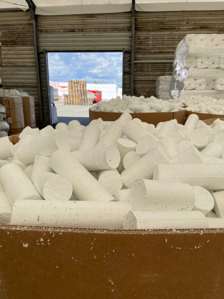 Eine Lagerhalle mit offener Laderampe, im Vordergrund sind mit Verpackungsmaterial aus Schaumstoff „Styropor“ gefüllte Kisten zu sehen, im Hintergrund Palettenstapel mit unterschiedlichen Waren.