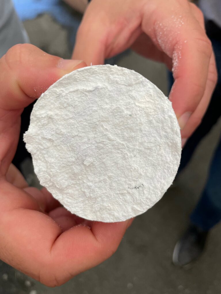 Eine Nahaufnahme einer Hand, die ein rundes Stück weißes Styropor hält, mit Fokus auf der rauen Oberfläche. Auf dem Material sind subtile Einkerbungen und ein kleiner dunkler Fleck sichtbar
