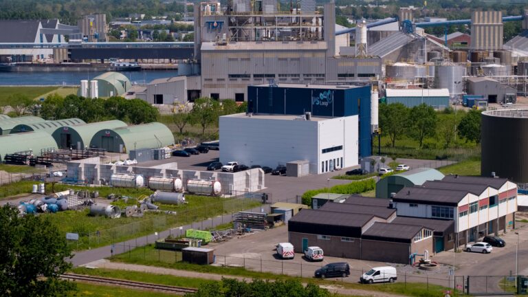 Luftaufnahme eines Industriegebiets mit mehreren Gebäuden, Lagertanks und geparkten Lastwagen in der Nähe einer Uferpromenade. Im Hintergrund sind nachhaltige Isoliermaterialien sichtbar.