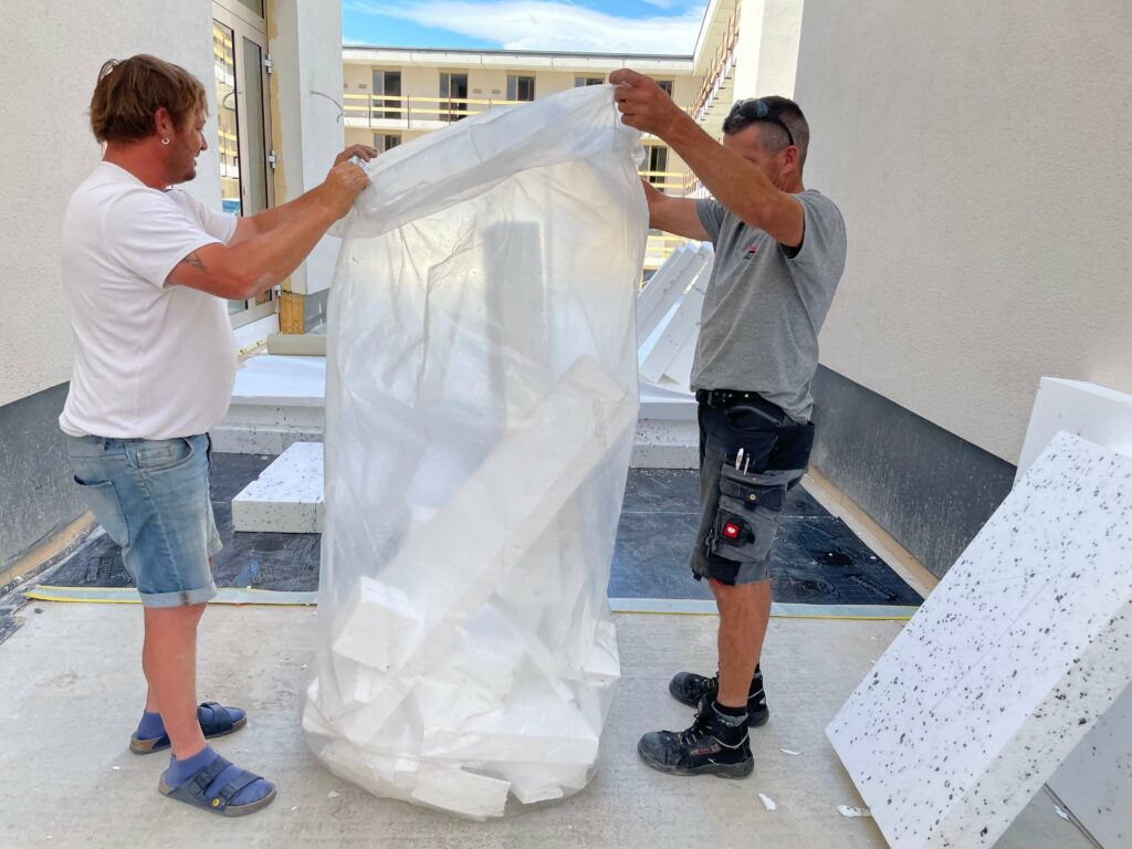 Zwei Bauarbeiter stehen zwischen Baumaterialien auf einer Baustelle. Einer hält eine große EPS-Hartschaumplatte, der andere hilft beim Auseinanderfalten.